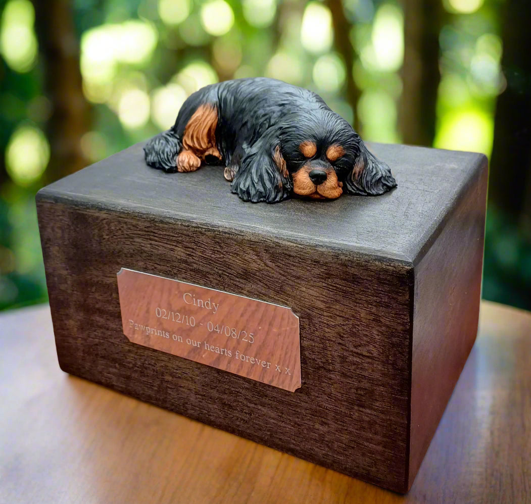 Wooden pet urn with a dog figurine on top, set against a blurred green outdoor background.